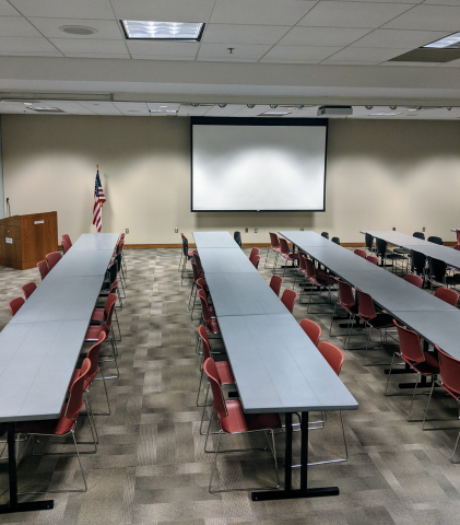 Rows of tables with chairs, a screen at the front of the room, and a podium on the left.